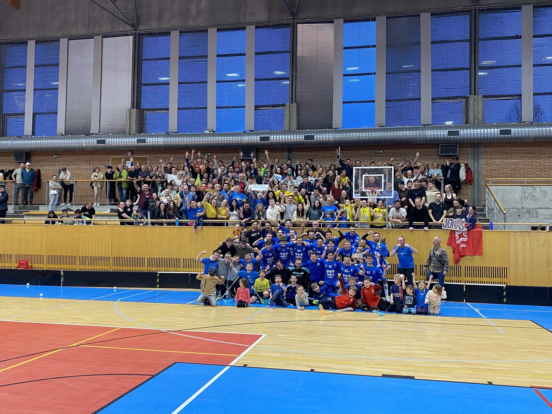Litomyšlští florbalisté slaví na palubovce postup; za nimi je plná tribuna fanoušků a na zábradlí visí červená vlajka s litomyšlskou lilií.