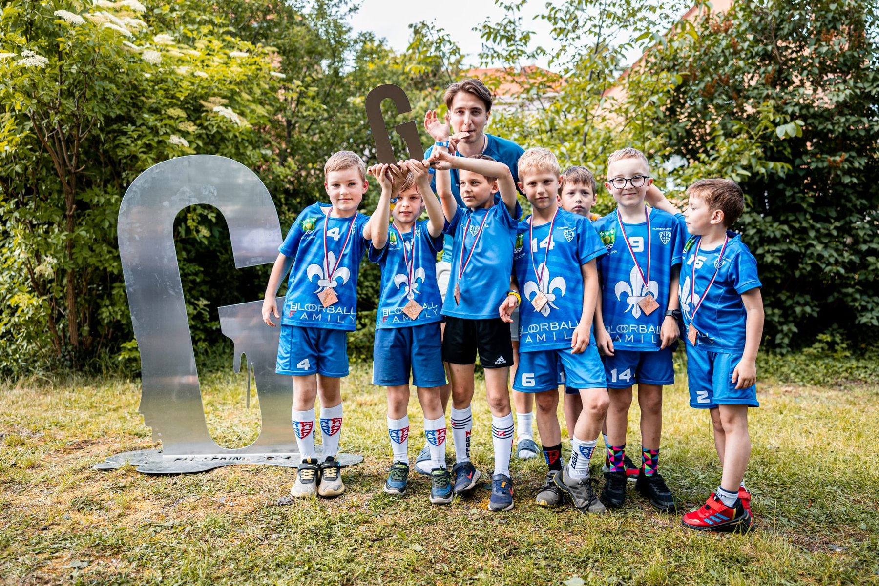 Nejmladší florbalisté s bronzovými medailemi na krku stojí v trávě vedle velkého průsvitného písmene G a společně zvedají nad hlavu kovovou trofej.