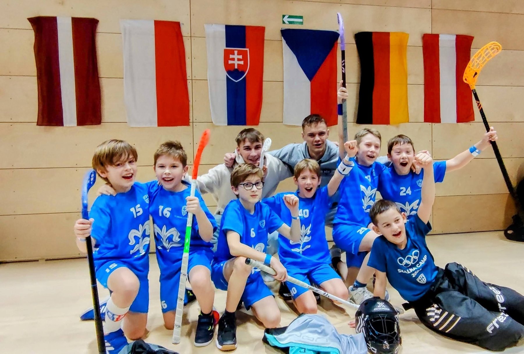 Litomyšlští florbalisté slaví v hale s holemi zdviženými nad hlavu. Na stěně za nimi visí vlajky Lotyšska, Polska, Slovenska, Česka, Německa a Rakouska.