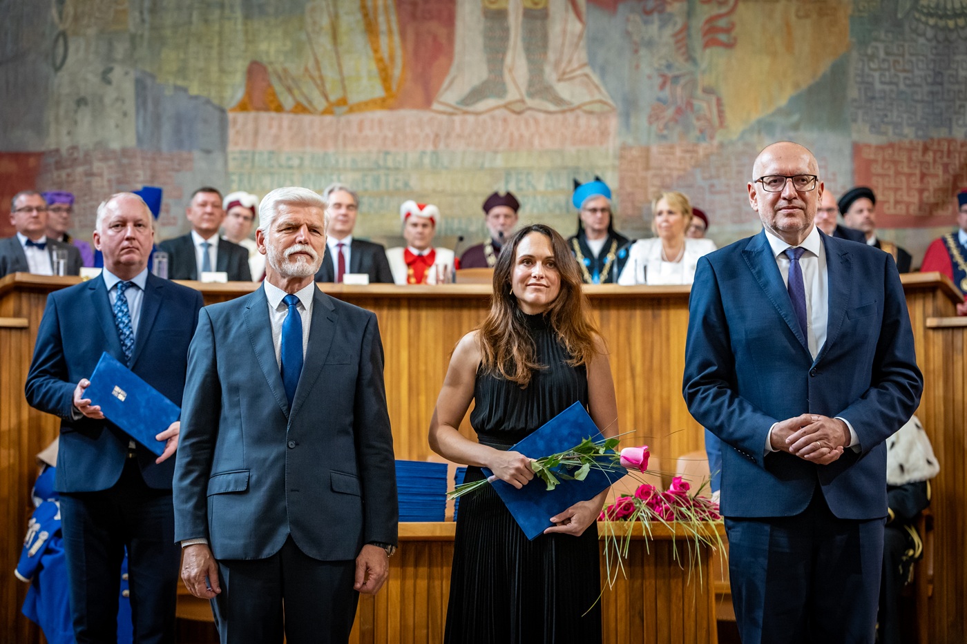 Zuzana Havránková stojí mezi prezidentem Petrem Pavlem a ministrem školství Mikulášem Bekem, v ruce drží modré desky se jmenovacím dekretem a kytici růží; v pozadí je freska Velké auly a řada dalších nově jmenovaných profesorů.