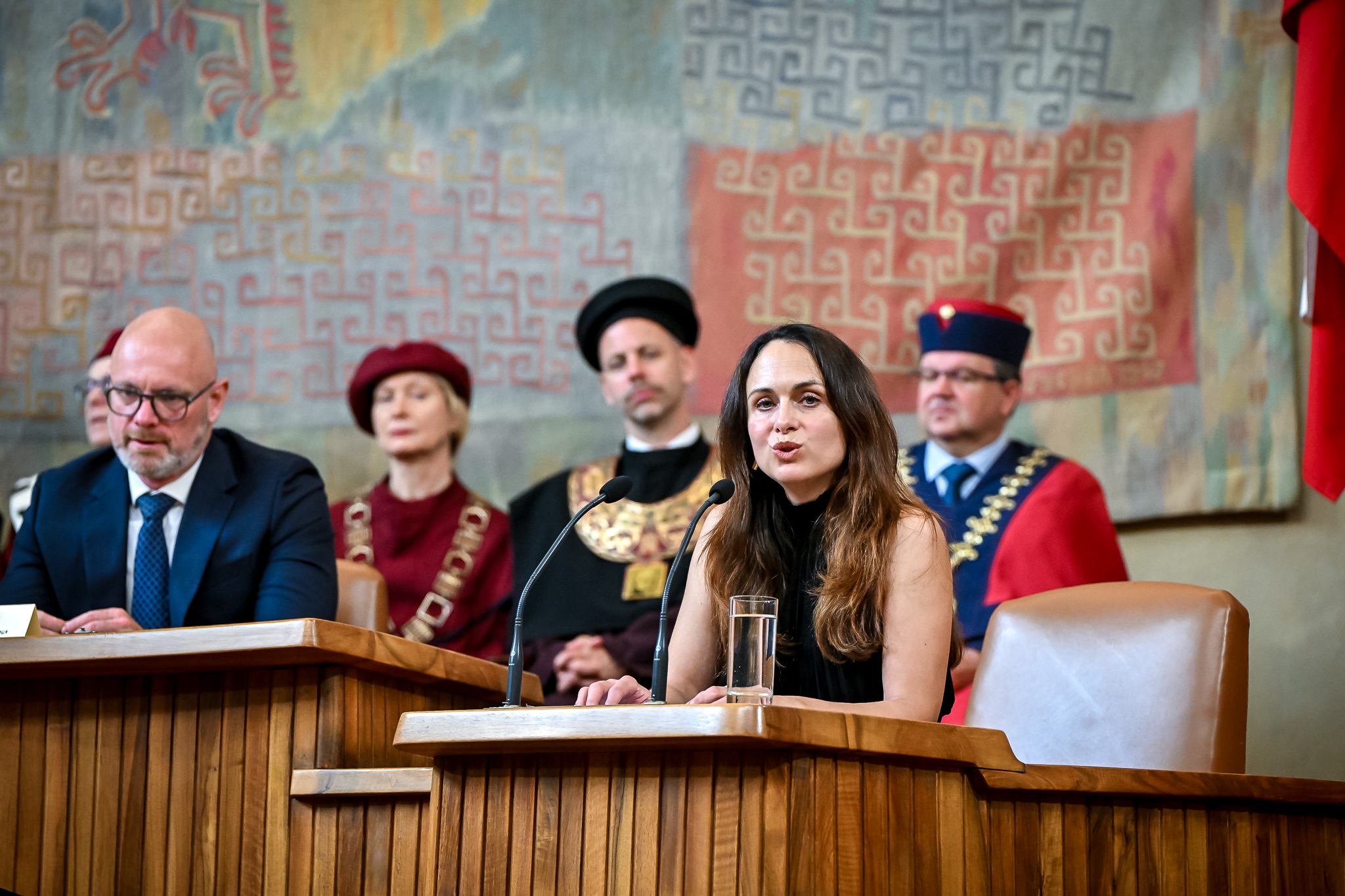 Zuzana Havránková mluví u řečnického pultu ve Velké aule Karolina; za ní sedí hosté ceremoniálu, část z nich v talárech s rektorskými řetězy, v pozadí visí gobelín.