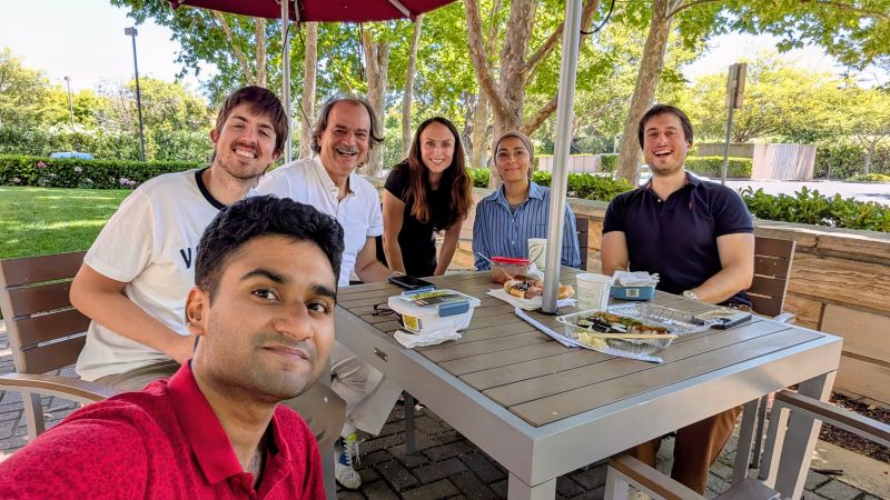 Group selfie of six people around an outdoor table on a sunny campus terrace, lunch containers and drinks on the table, trees and a lawn behind them.
